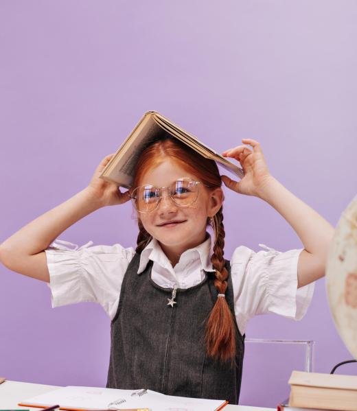 Funny red haired girl with freckles and stylish hairstyle in school blouse and grey sundress holding book on her head and smiling..